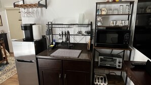 Eine kompakte Küche mit Kühlschrank, Mikrowelle und einer Kaffeemaschine. Ein Regal mit Glasware und ein Hängegestell für Besteck sind vorhanden.