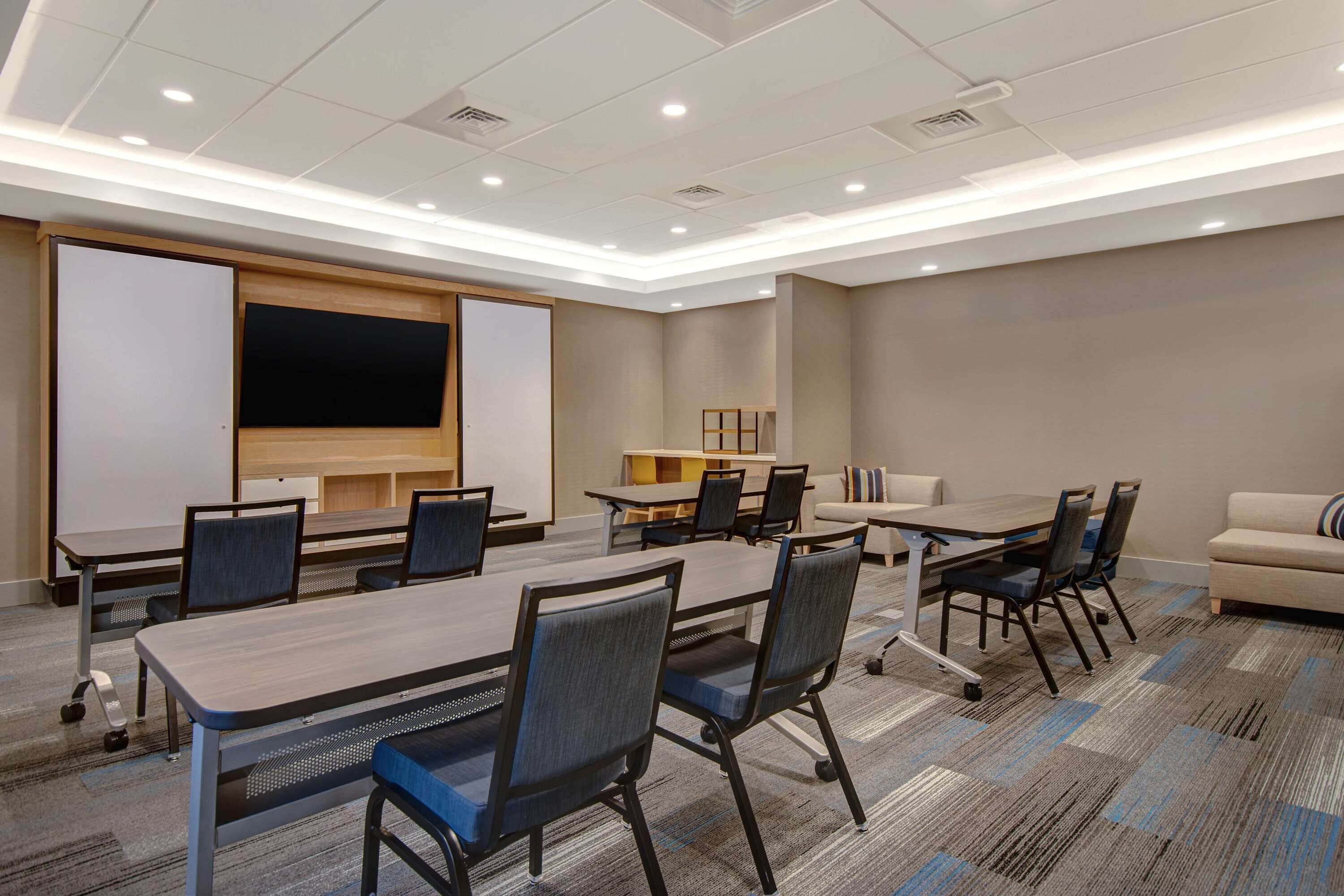 Un salón de conferencias con una gran mesa, sillas, una pantalla de proyector, un sofá y una pequeña mesa con sillas.