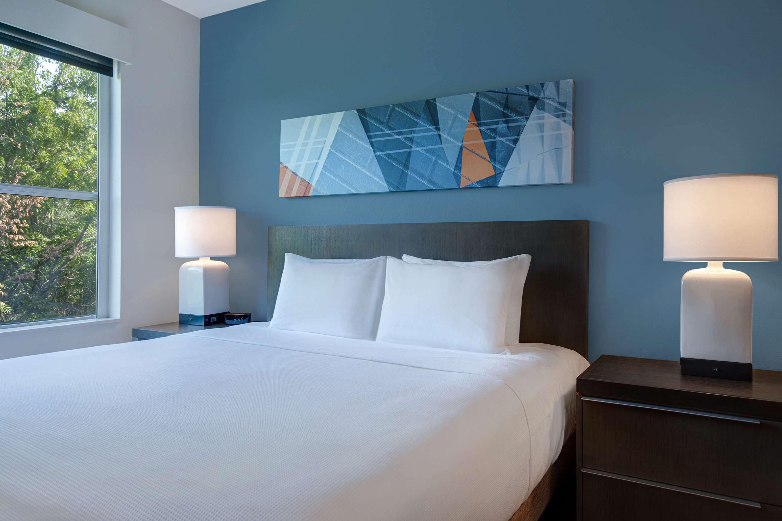 Habitación de hotel con una cama grande, dos lámparas de noche, una mesita de noche y un cuadro abstracto colgado en la pared.