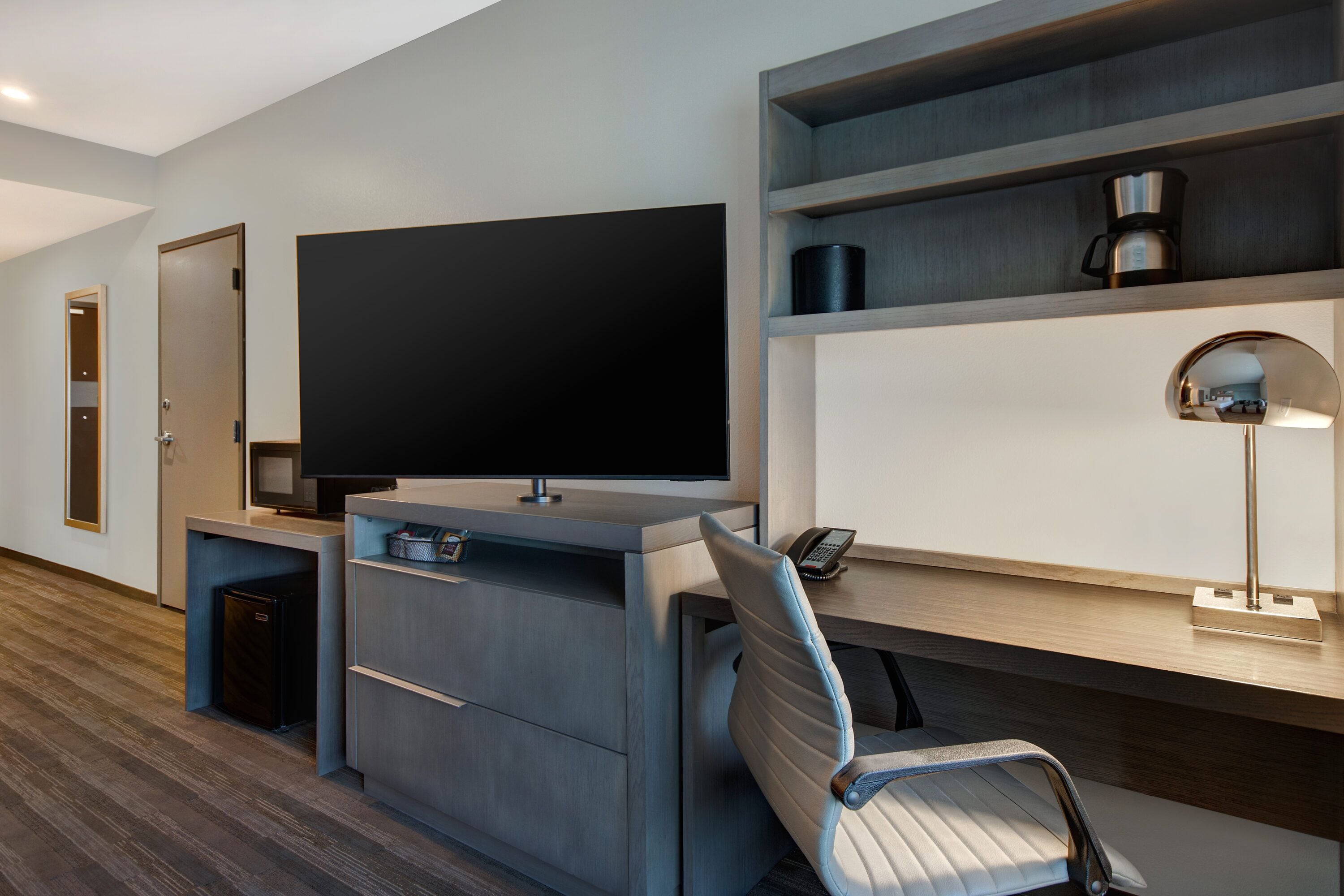 A modern hotel room with a large flat-screen TV, a desk with a lamp, a chair, and a coffee machine on a shelf.