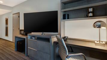 A modern hotel room with a large flat-screen TV, a desk with a lamp, a chair, and a coffee machine on a shelf.