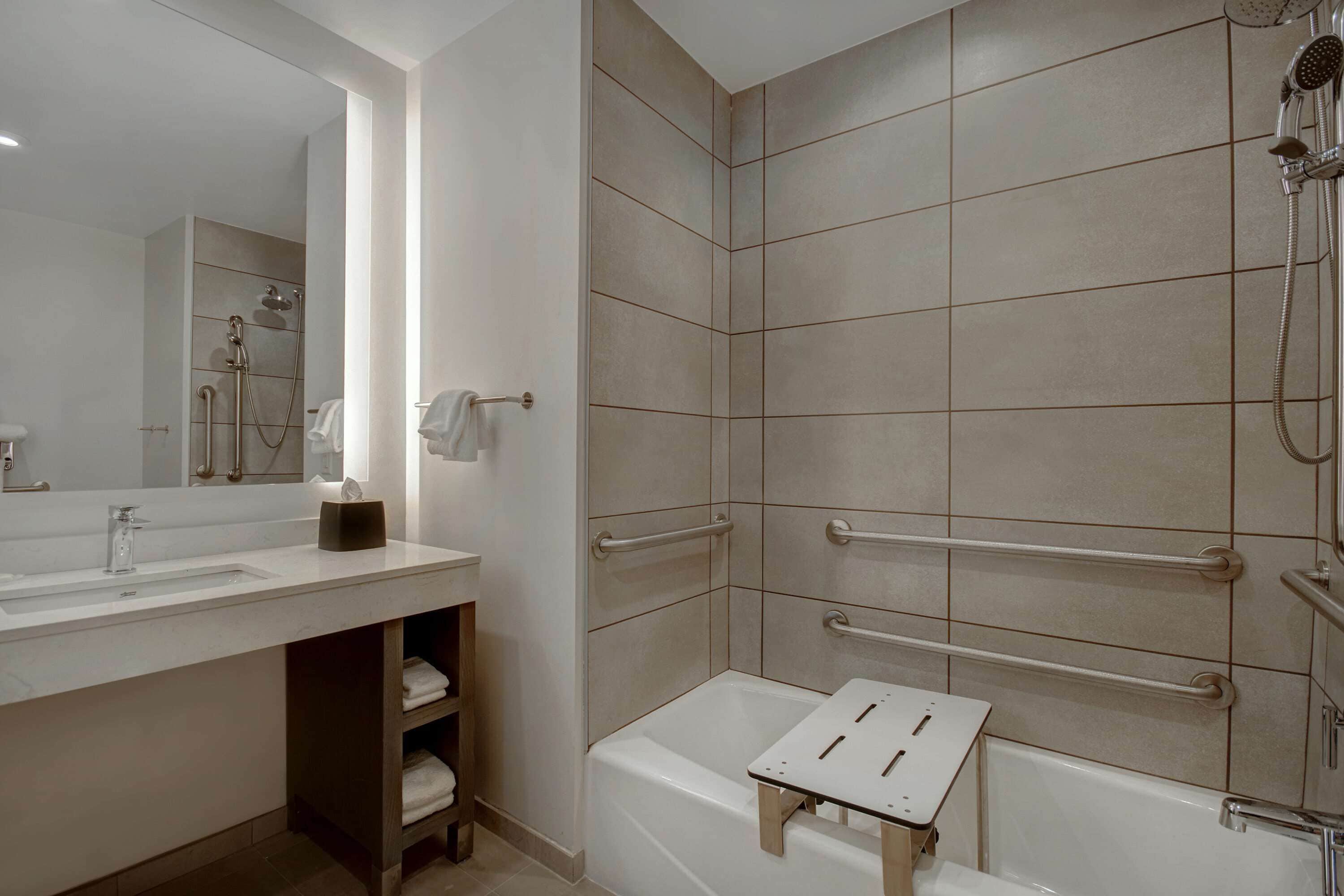 A modern bathroom with a large mirror, a sink, a shower with grab bars, and a bathtub.