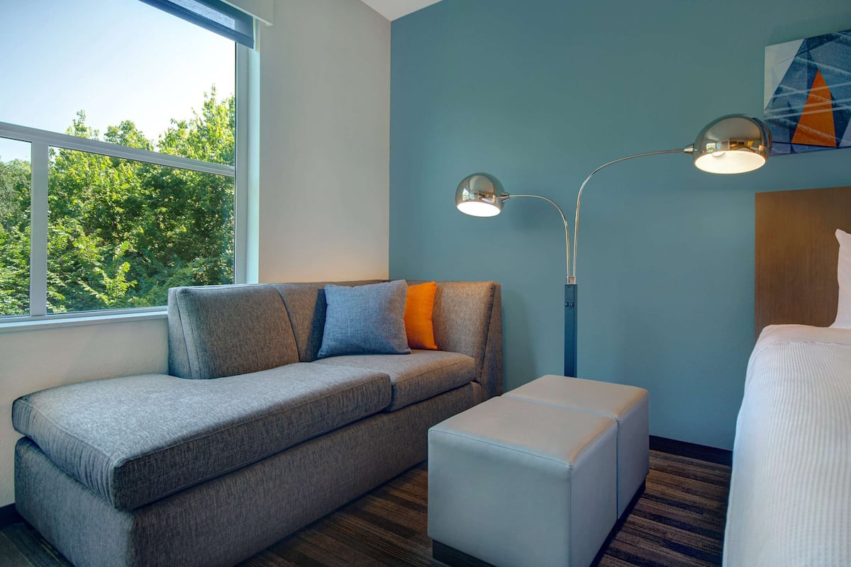 Une chambre d’hôtel moderne avec un canapé gris, un pouf blanc et une grande fenêtre donnant sur des arbres.