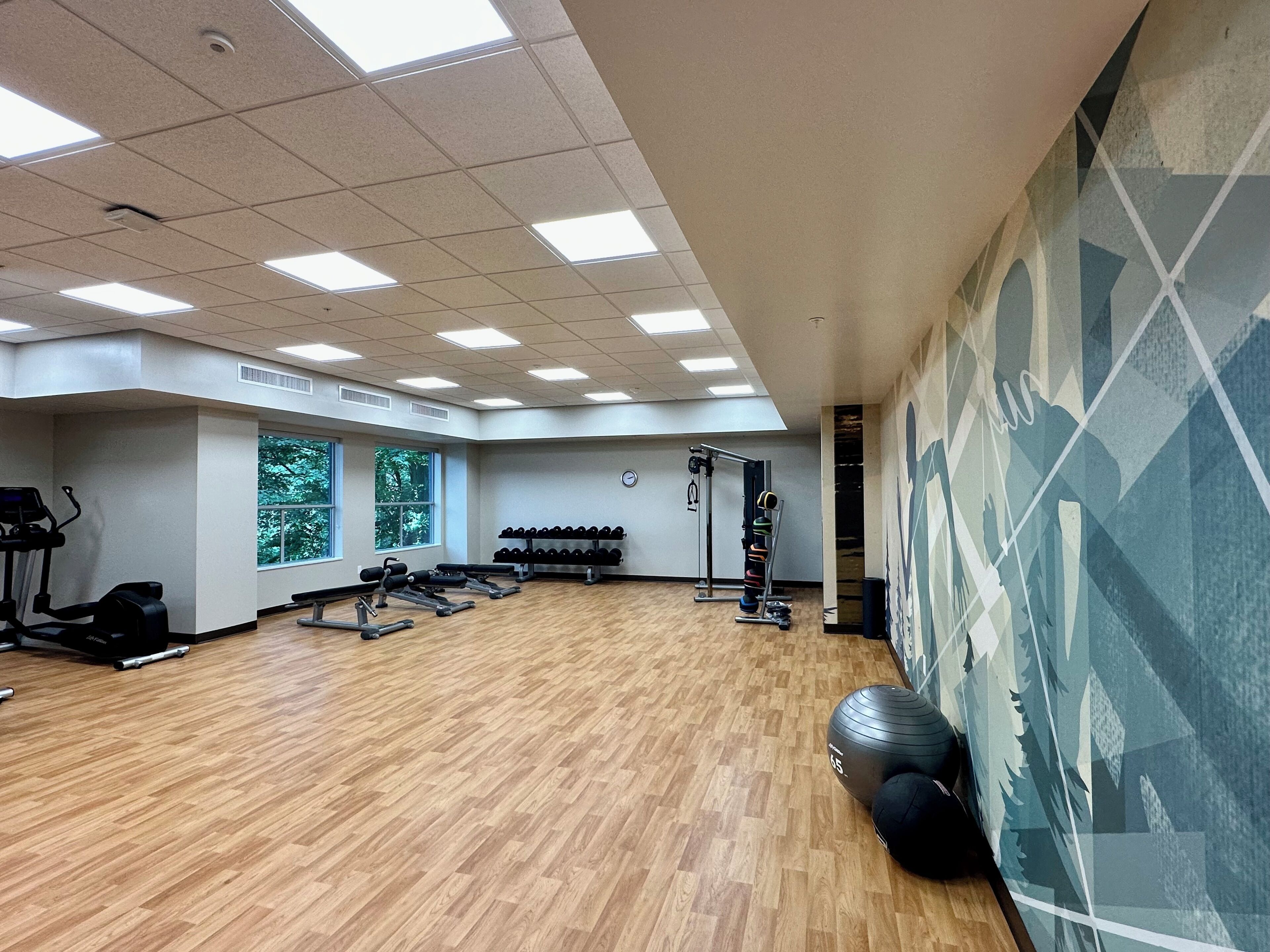 A spacious gym with wooden flooring, various exercise equipment, and large windows providing natural light.