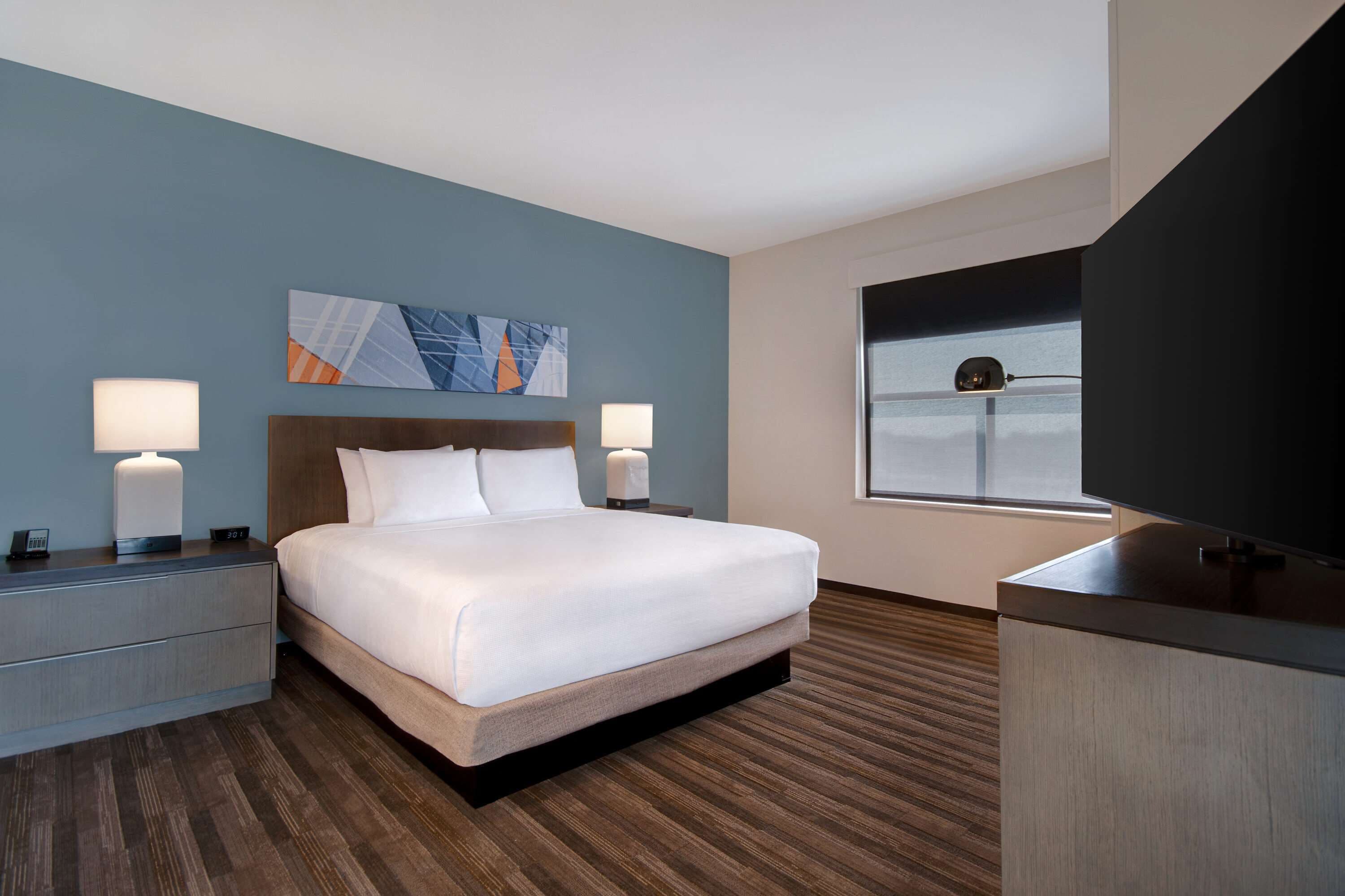 A modern hotel room with a large bed, two bedside tables with lamps, a flat-screen TV, and a window with a view.