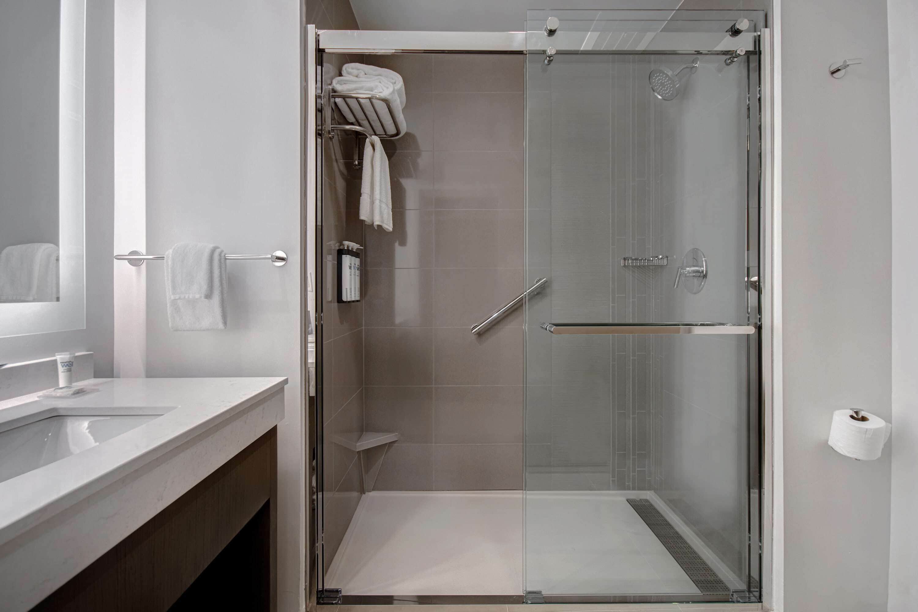 Un baño moderno con ducha envuelta en vidrio, un lavamanos blanco y un perchero con toallas dobladas.