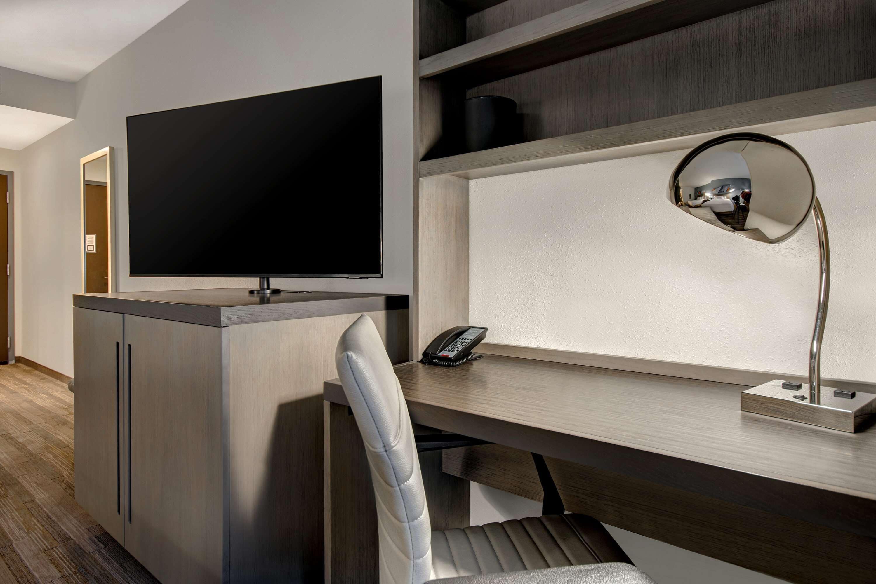 A modern hotel room with a flat-screen TV mounted on a cabinet, a desk with a telephone and a mirror, and a cushioned chair.