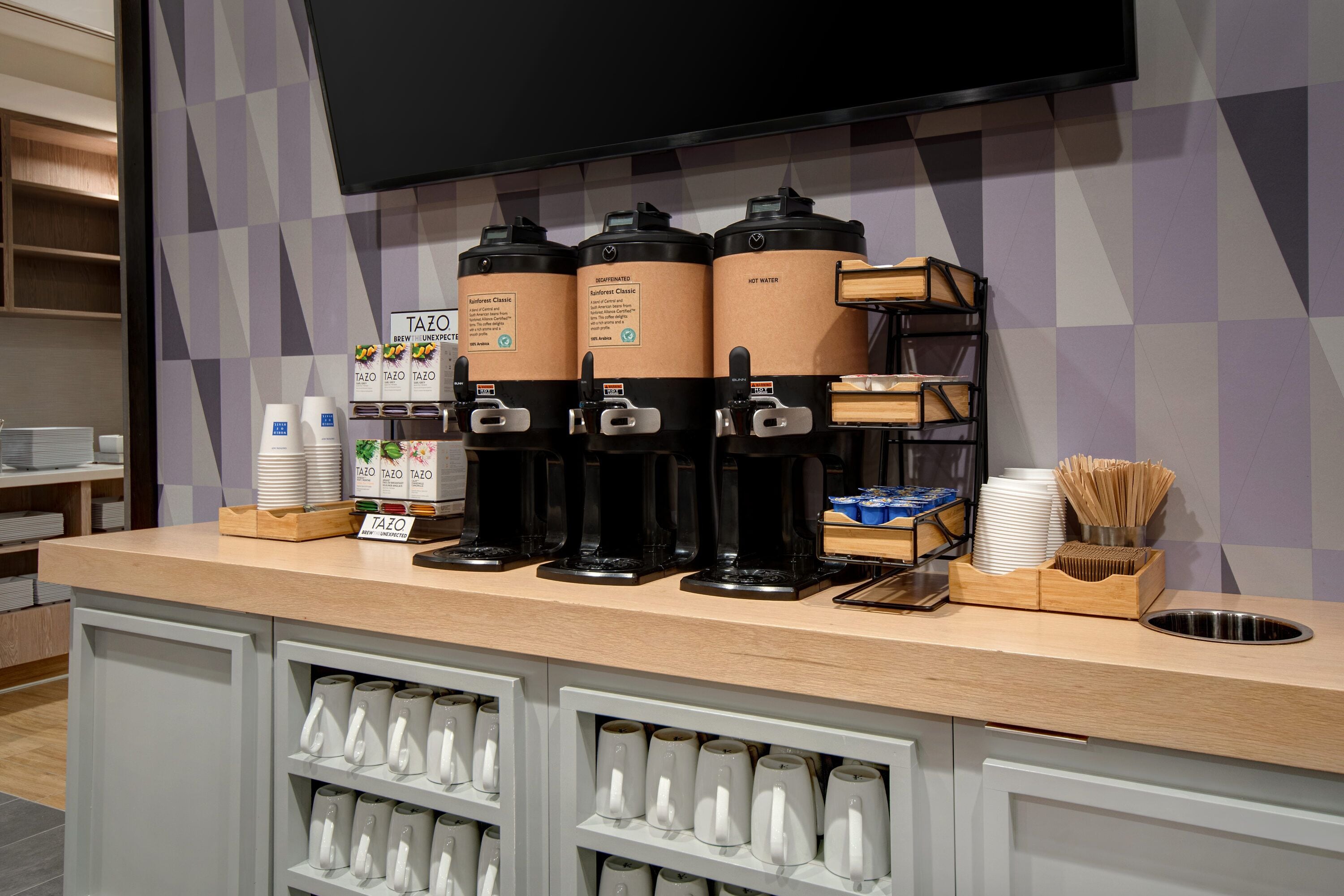 Eine Kaffeestation mit drei Spendern, beschriftet mit 'Tazo' und 'Warmes Wasser', einer Stapel weißer Tassen und einer Holztablett mit Essstäbchen und einer Schüssel.