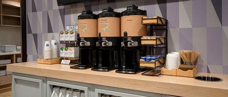 Eine Kaffeestation mit drei Spendern, beschriftet mit 'Tazo' und 'Warmes Wasser', einer Stapel weißer Tassen und einer Holztablett mit Essstäbchen und einer Schüssel.