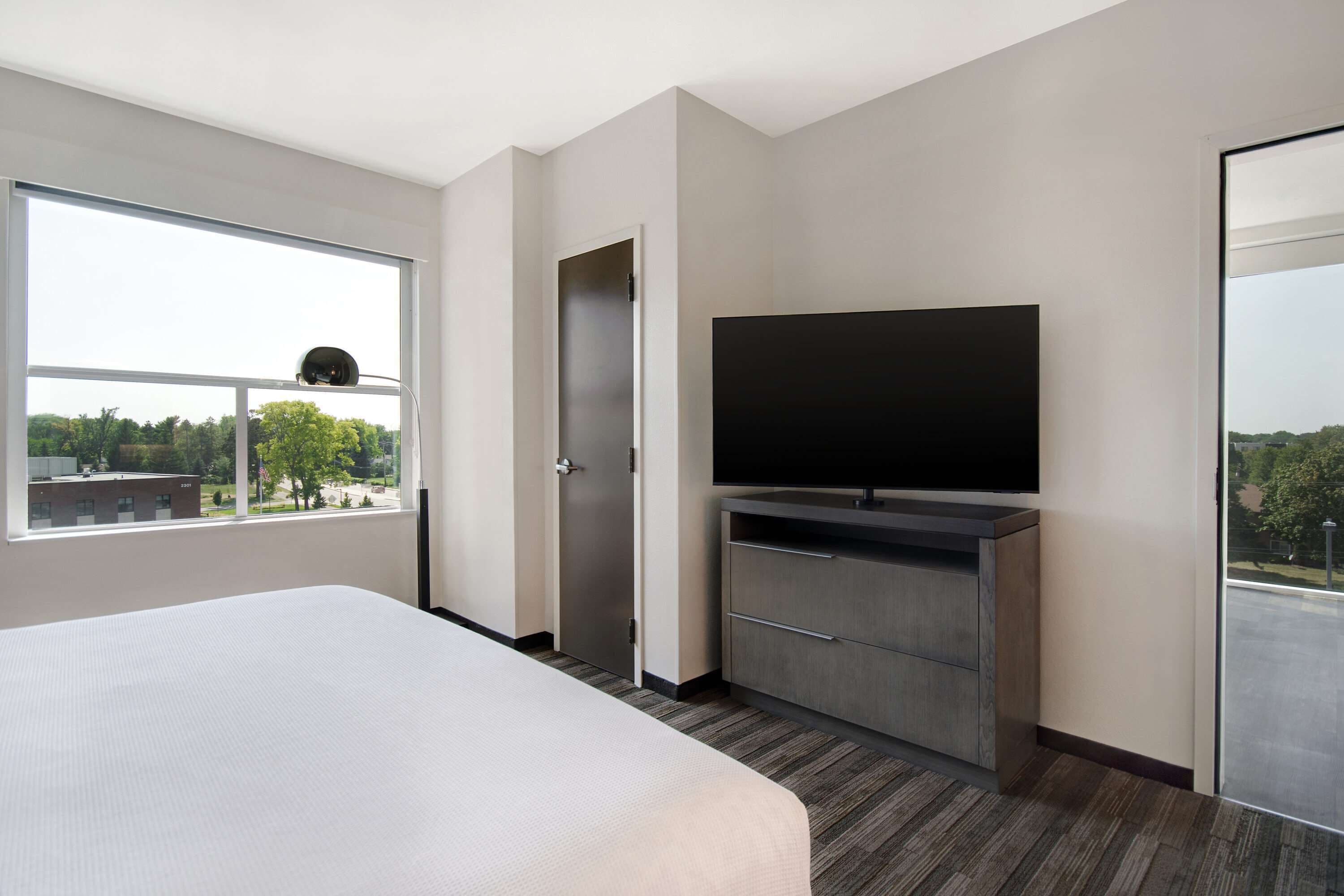 A modern hotel room with a large bed, a flat-screen TV, a wardrobe, and a window offering a city view.