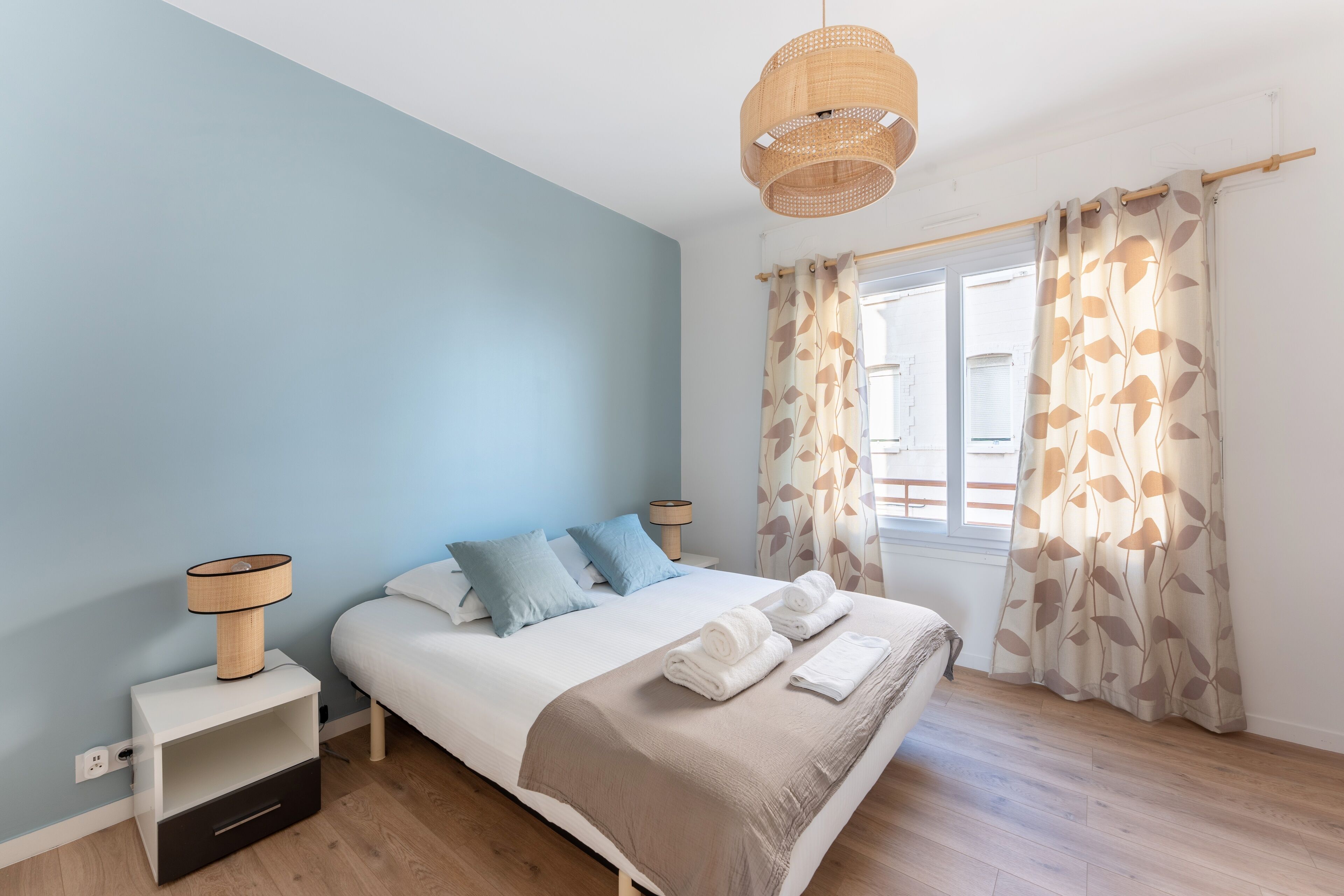 A bedroom with a bed, bedside tables, a lamp, a window with curtains, and a wooden floor.