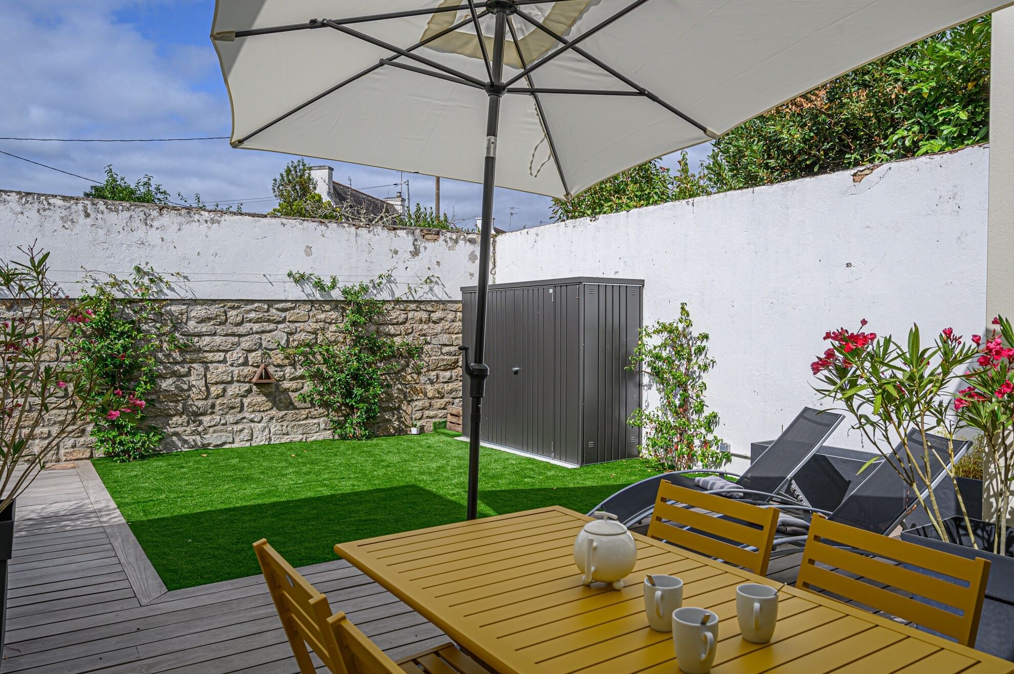 Une terrasse avec une table et des chaises en bois, un parasol blanc et un mur en pierre.