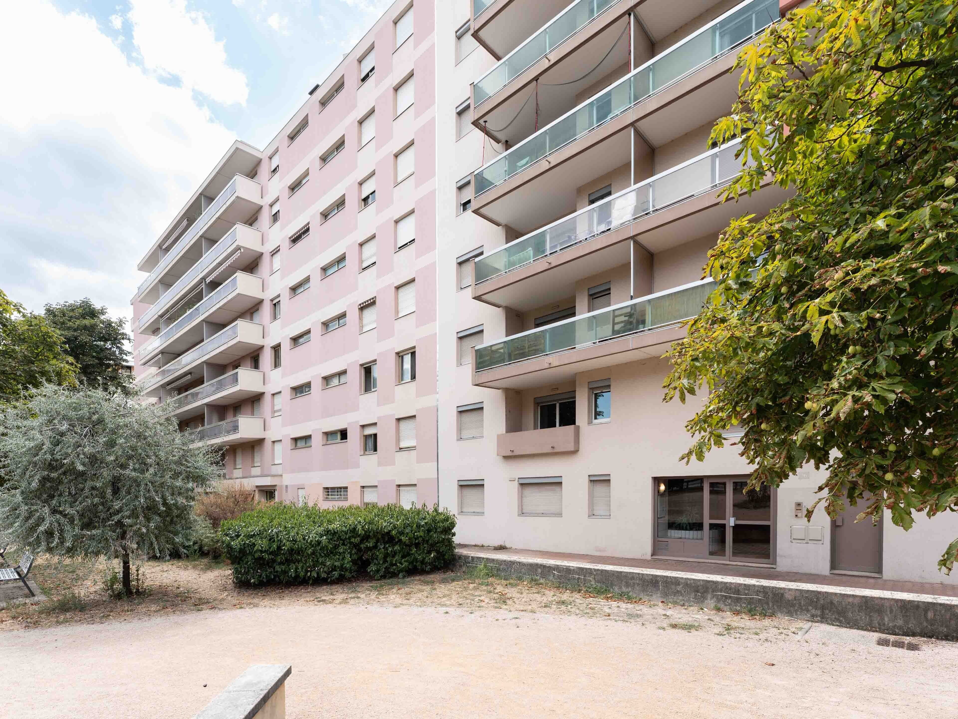 A multi-story residential building with balconies, a paved area, and a tree.