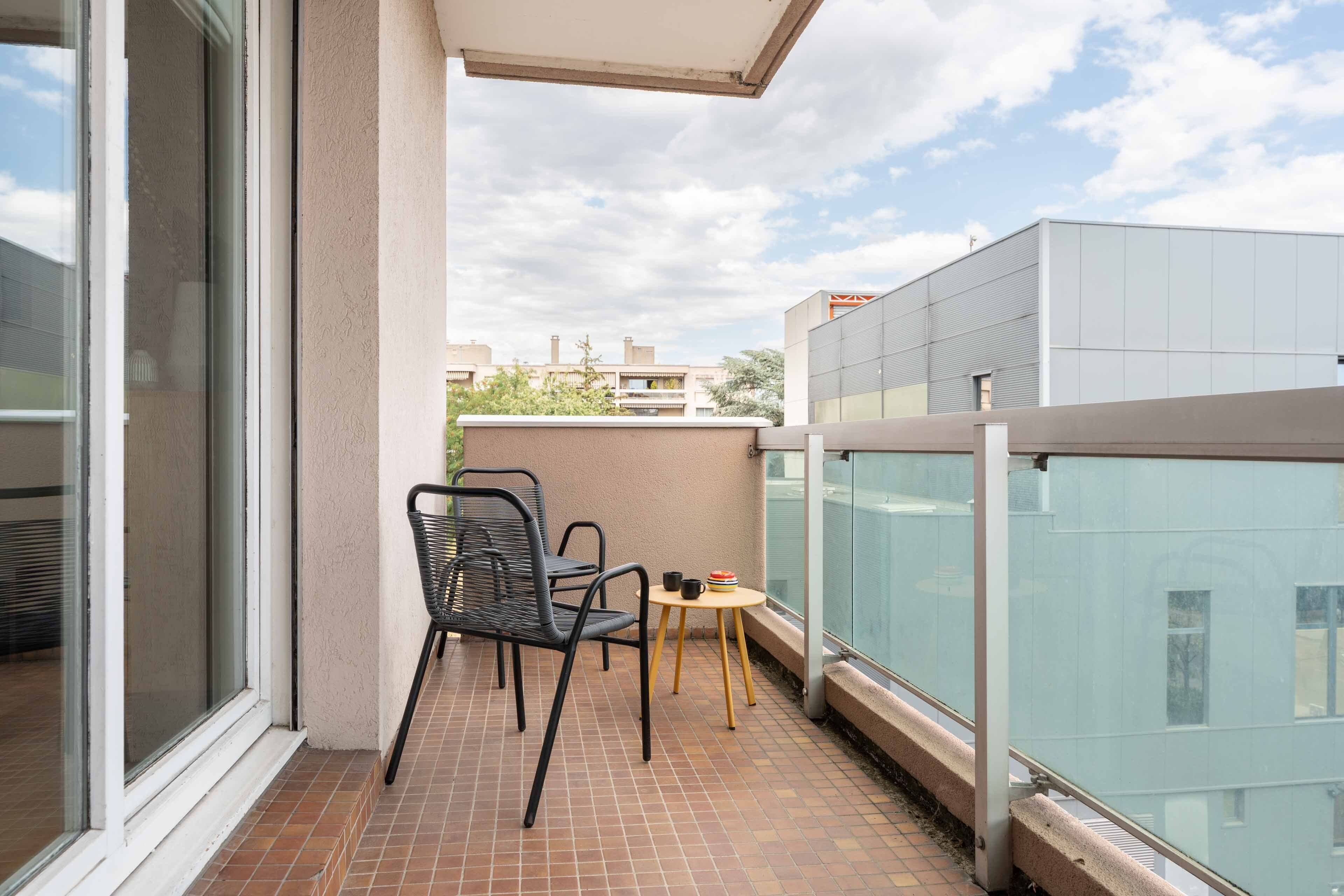 A balcony with two chairs and a small table, a glass railing, and a view of buildings in the background.