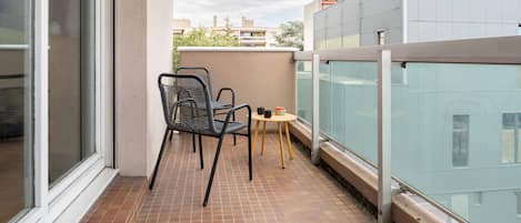 Ein Balkon mit zwei Stühlen und einem kleinen Tisch, einer Glasreling und Blick auf Gebäude im Hintergrund.