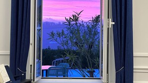 Una habitación con ventana que ofrece vistas de un atardecer sobre el agua, cortinas y un piano.