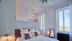Un dormitorio con una cama grande, una mesita de noche con lámpara, un espejo y vistas al exterior a través de una ventana.