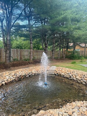 A fountain surrounded by a pebble border, with trees and a wooden fence in the background.
