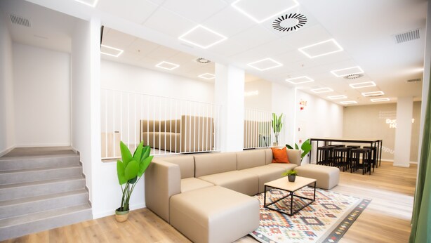 A modern living room with a beige sectional sofa, a coffee table, and a patterned rug. There is a staircase on the left and a dining area with tables and chairs in the background. - Micampus Residencia Estudiantes Burjassot Parque (Burjasot)