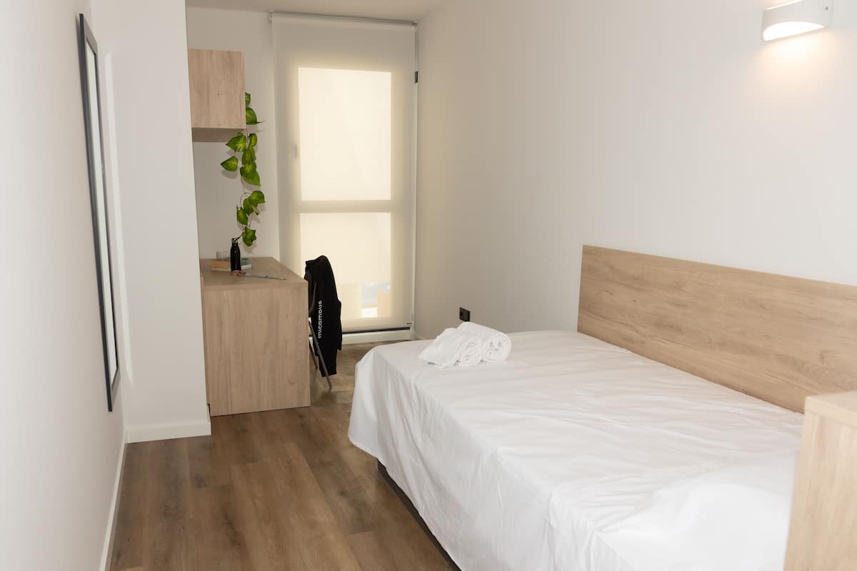 A modern hotel room with a wooden headboard, a neatly made bed, a small desk with a plant, and a sliding door leading to a bright room.