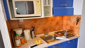 Cocina compacta con microondas, pileta y muebles azules. El salpicadero tiene baldosas naranjas, una tabla de cortar con un cuchillo y una botella de jugo de naranja.