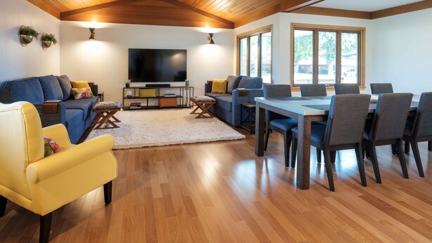Ein modernes Wohnzimmer mit Holzboden, einem Flachbildfernseher, einer Couch, einem Esstisch mit Stühlen und großen Fenstern.