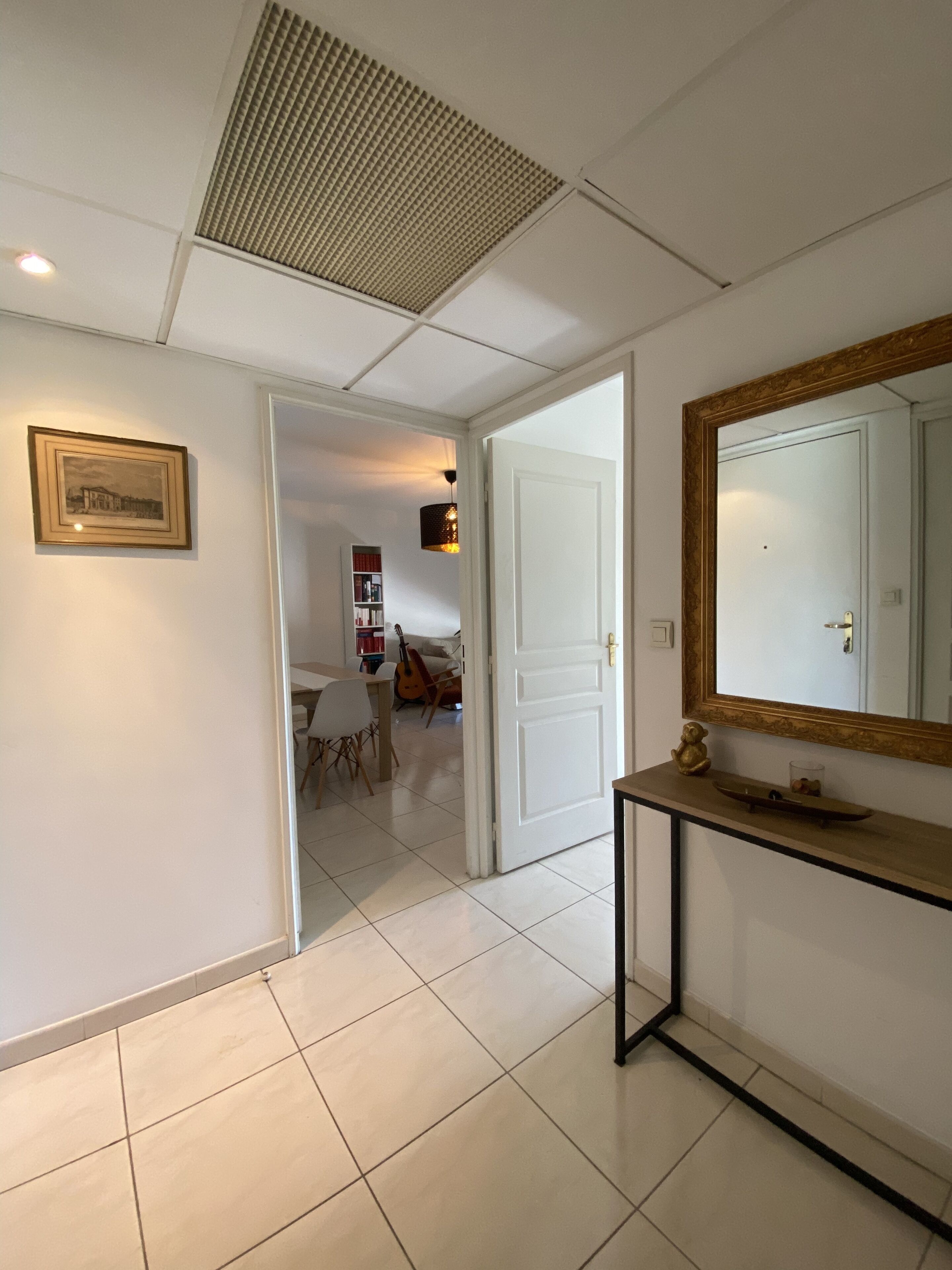 A hallway with a tiled floor, a framed picture on the wall, a white door, and a console table with a mirror and a small shelf.