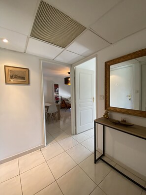Un pasillo con suelo de baldosas, un cuadro en la pared, una puerta blanca y una mesa consola con espejo y un estante pequeño.