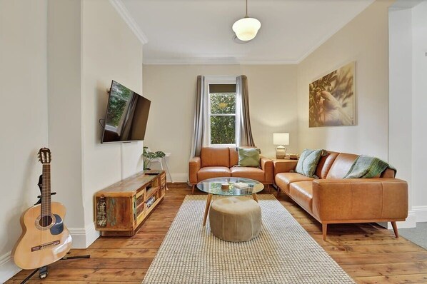 A living room with a sofa, ottoman, and a glass coffee table. - Best Nest on March. Firepit Cottage. Walk to CBD. (Orange)