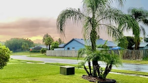 Un barrio residencial con un césped cuidado, una palmera y una casa azul con cerca de madera.