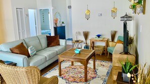 Una sala de estar con un sofá azul claro, una mesa de centro de madera, sillas de mimbre, una alfombra estampada y un mueble de televisión con una pantalla plana.