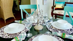 Mesa de comedor dispuesta con platos blancos, vasos y un centro de mesa con velas, rodeada de sillas de mimbre y un adorno de pared.