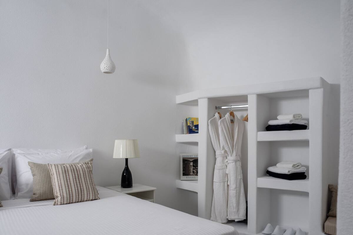 A modern bedroom with a large bed, a bedside table with a lamp, a robe rack, and built-in shelves with folded towels and shoes.
