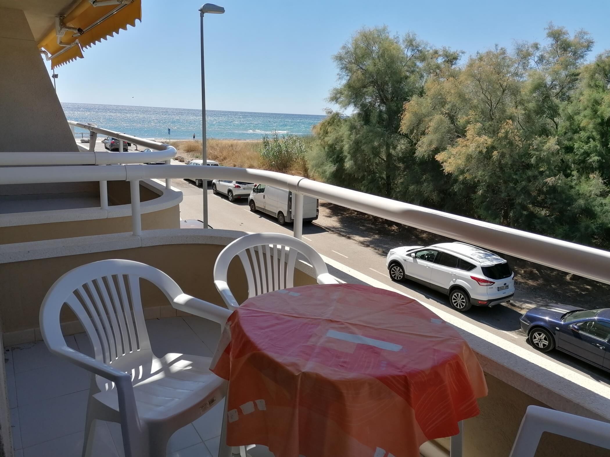 A balcony with white plastic chairs and a small table, overlooking a road with parked cars and a view of the sea and trees.
