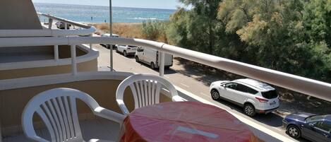 A balcony with white plastic chairs and a small table, overlooking a road with parked cars and a view of the sea and trees.