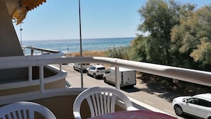 Un balcon avec vue sur la mer, une rue bordée de voitures garées et une voiture blanche au premier plan.
