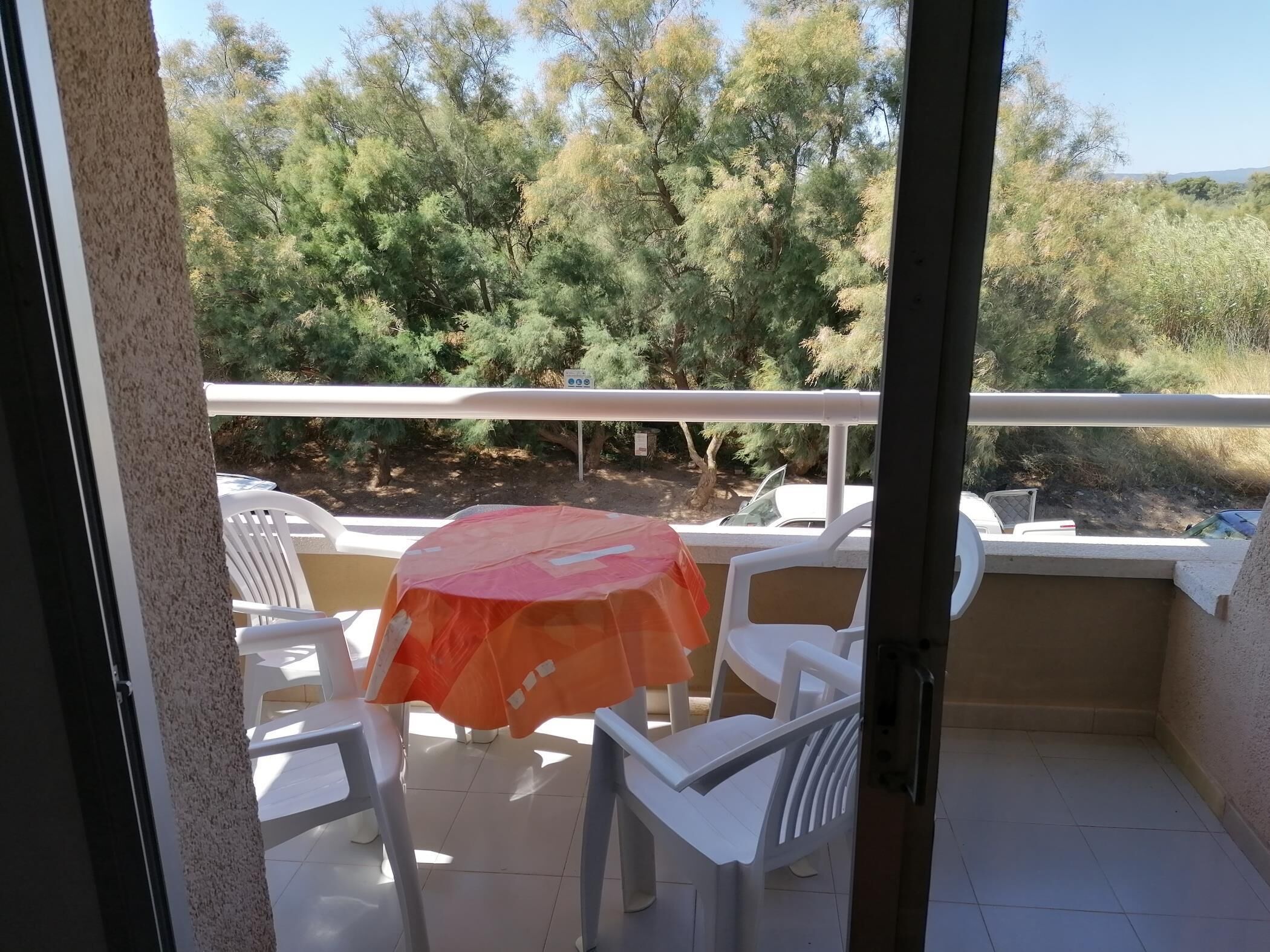 A balcony with white plastic chairs and a table, covered by an orange umbrella.