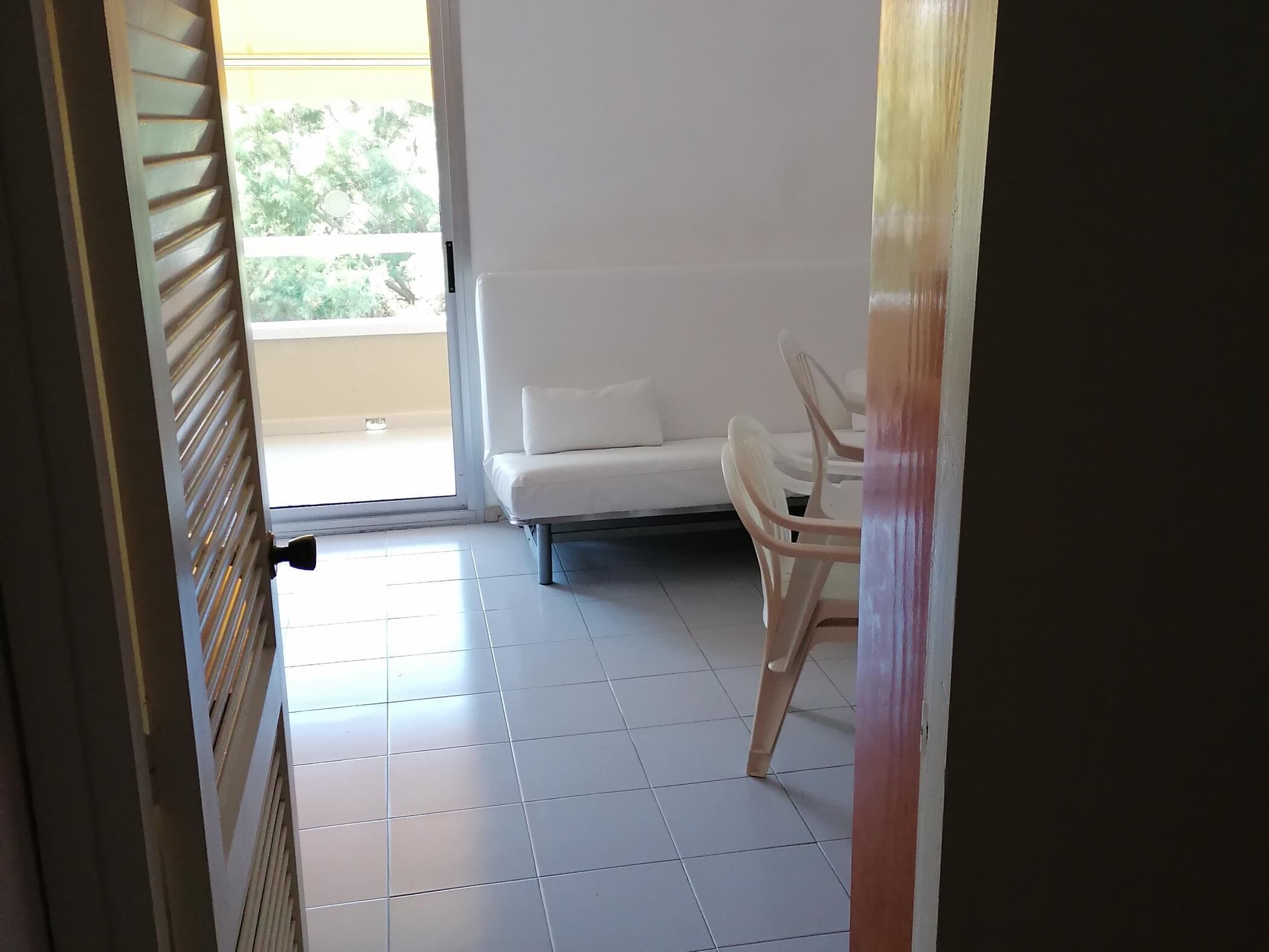 Une pièce avec un canapé blanc et un fauteuil, un sol carrelé et une porte donnant sur l’extérieur.