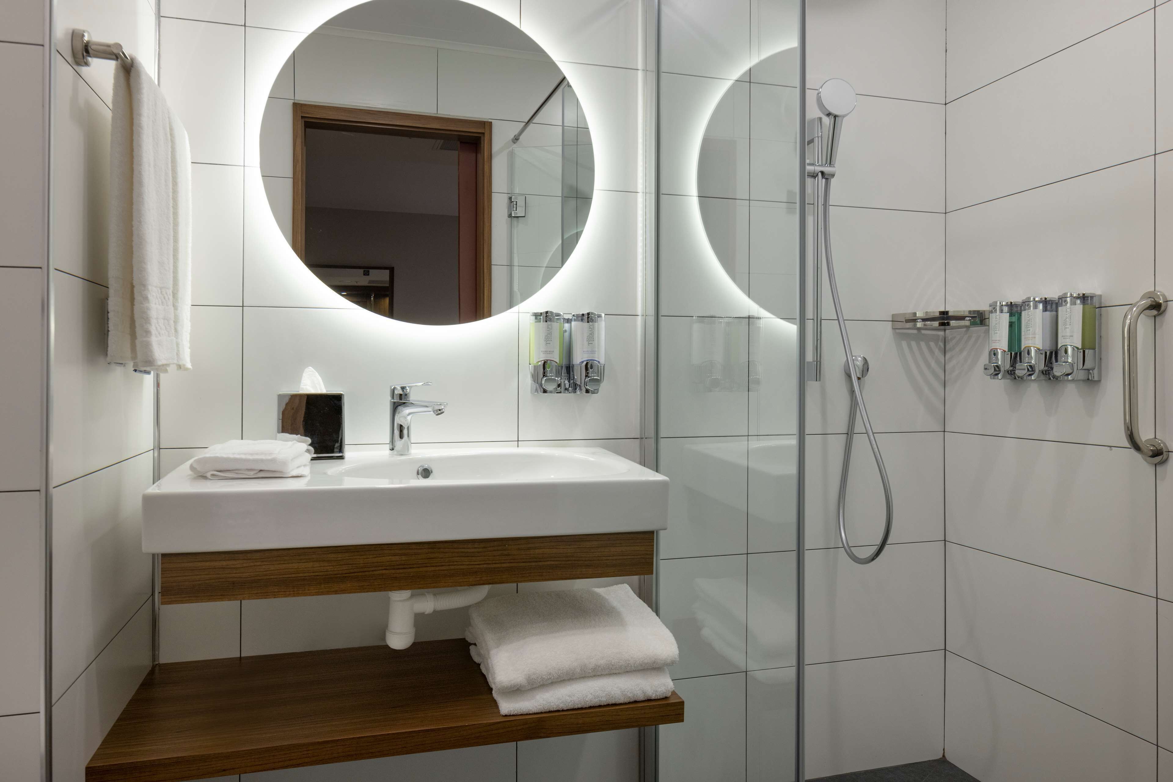 Casa de banho moderna com lavatório branco, espelho com luz circular, chuveiro e prateleira com toalhas.