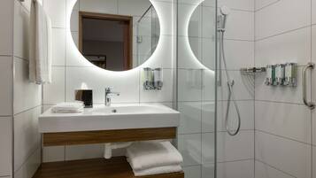 Casa de banho moderna com lavatório branco, espelho com luz circular, chuveiro e prateleira com toalhas.