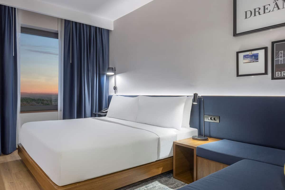 Ein Hotelzimmer mit Bett, Schreibtisch, Stuhl, Nachttisch mit Lampe und Blick auf den Sonnenuntergang durch das Fenster.