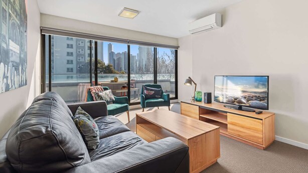 A modern living room with a leather sofa, a wooden coffee table, a teal armchair, and a TV on a wooden stand. - Chic Inner City Convenience, with Secure Parking (South Melbourne)
