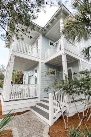 A two-story house with a white porch, a balcony, and a small front yard with plants.