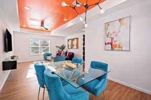 A modern living room with a glass dining table, blue chairs, a ceiling fan, and a large abstract painting.