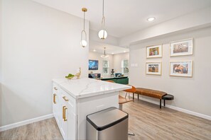 A modern kitchen with white cabinetry, a marble countertop, and pendant lighting. There is a bench along the wall and framed artwork on the wall.