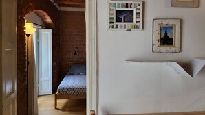Une pièce avec un lit, un plancher en bois, un mur en briques et une porte blanche. Deux tableaux encadrés sont accrochés au mur.