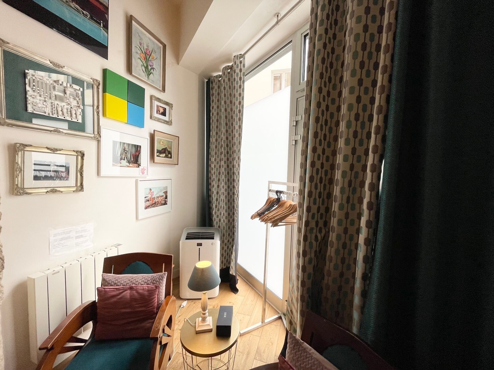 A compact room with a bed, a small table with a lamp, a radiator, and a window with curtains.