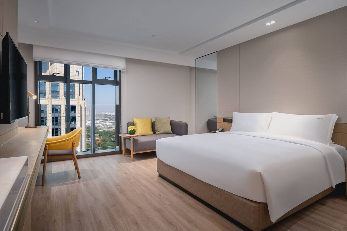 Habitación de hotel moderna con una cama grande, un escritorio con silla, un sofá y un amplio ventanal con vistas a la ciudad.