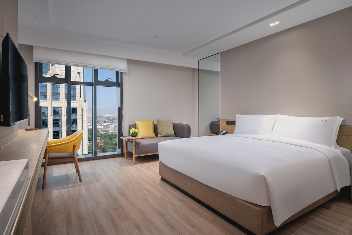 Habitación de hotel moderna con una cama grande, un escritorio con silla, un sofá y vistas a la ciudad a través de amplios ventanales.