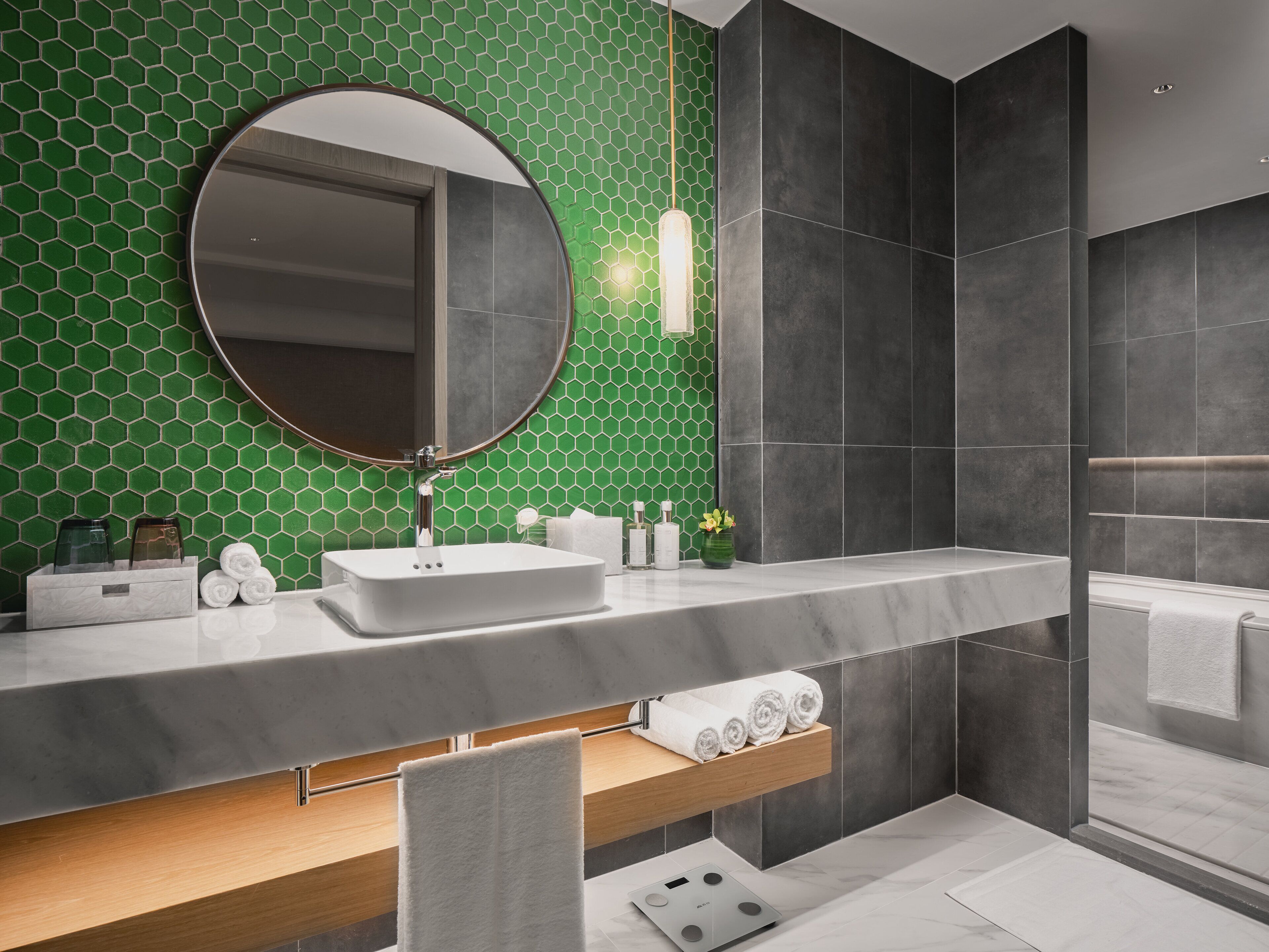 A modern bathroom with a green hexagonal tile backsplash, a round mirror, a white sink, and a wooden vanity.